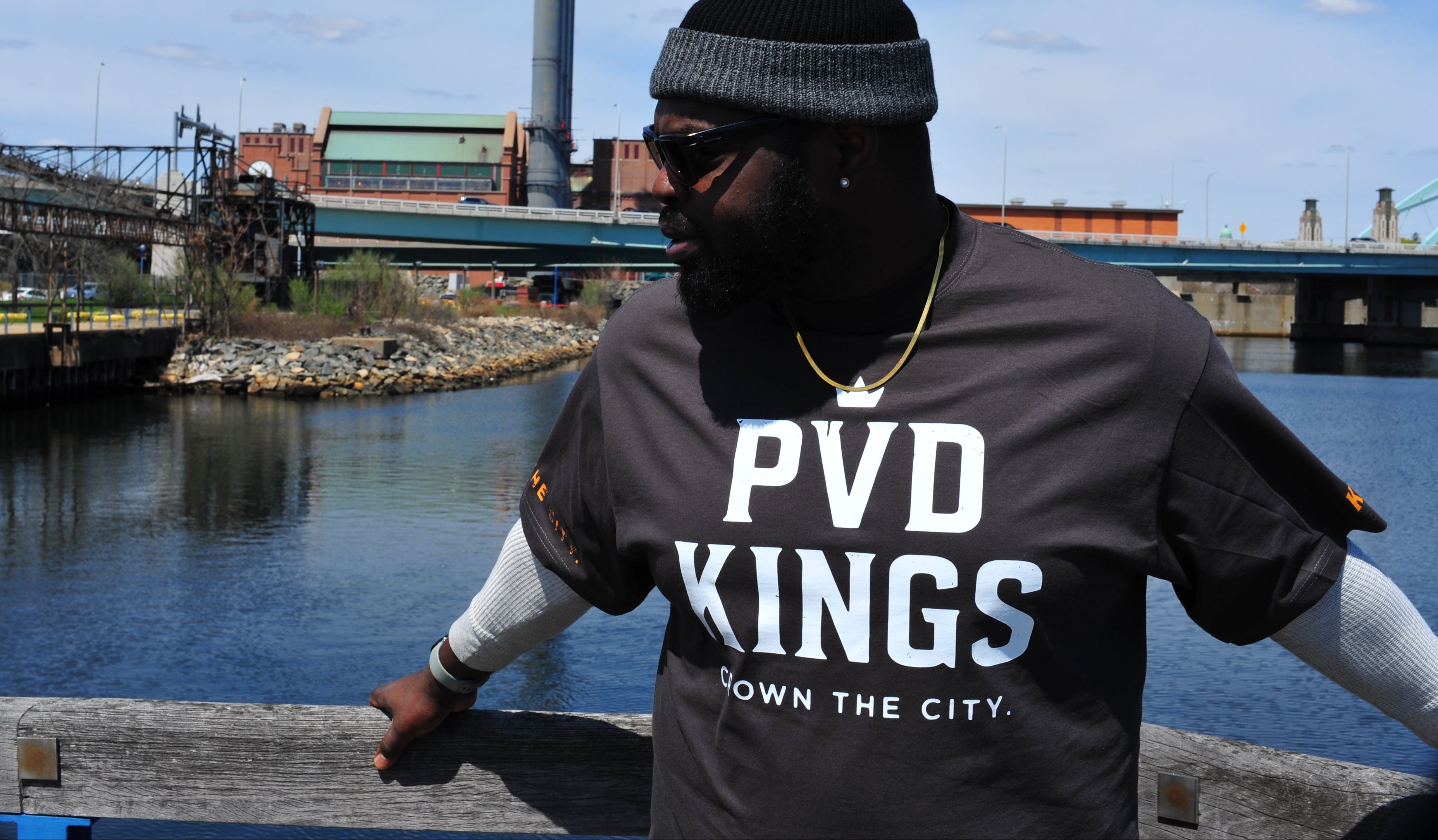 Mikefabz wearing a 'PVD Kings' shirt by a waterfront with industrial background