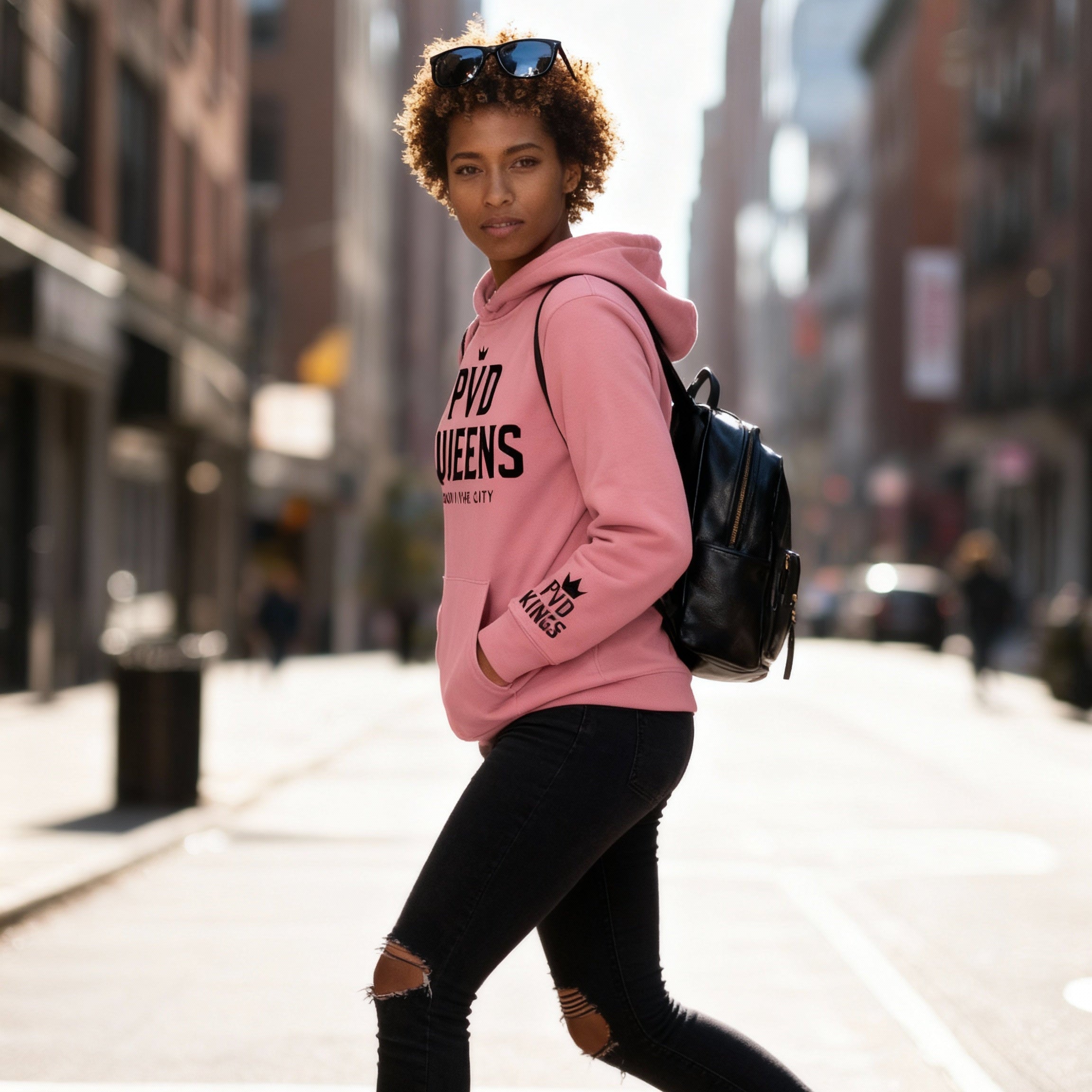 Person wearing a pink hoodie with text, black pants, and black shoes on a city street.